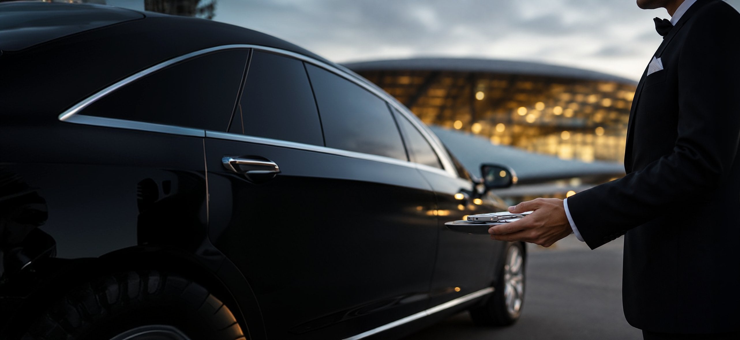 A chauffeur in a dark suit holds a tray with car keys beside a black sedan outside an airport terminal at dusk